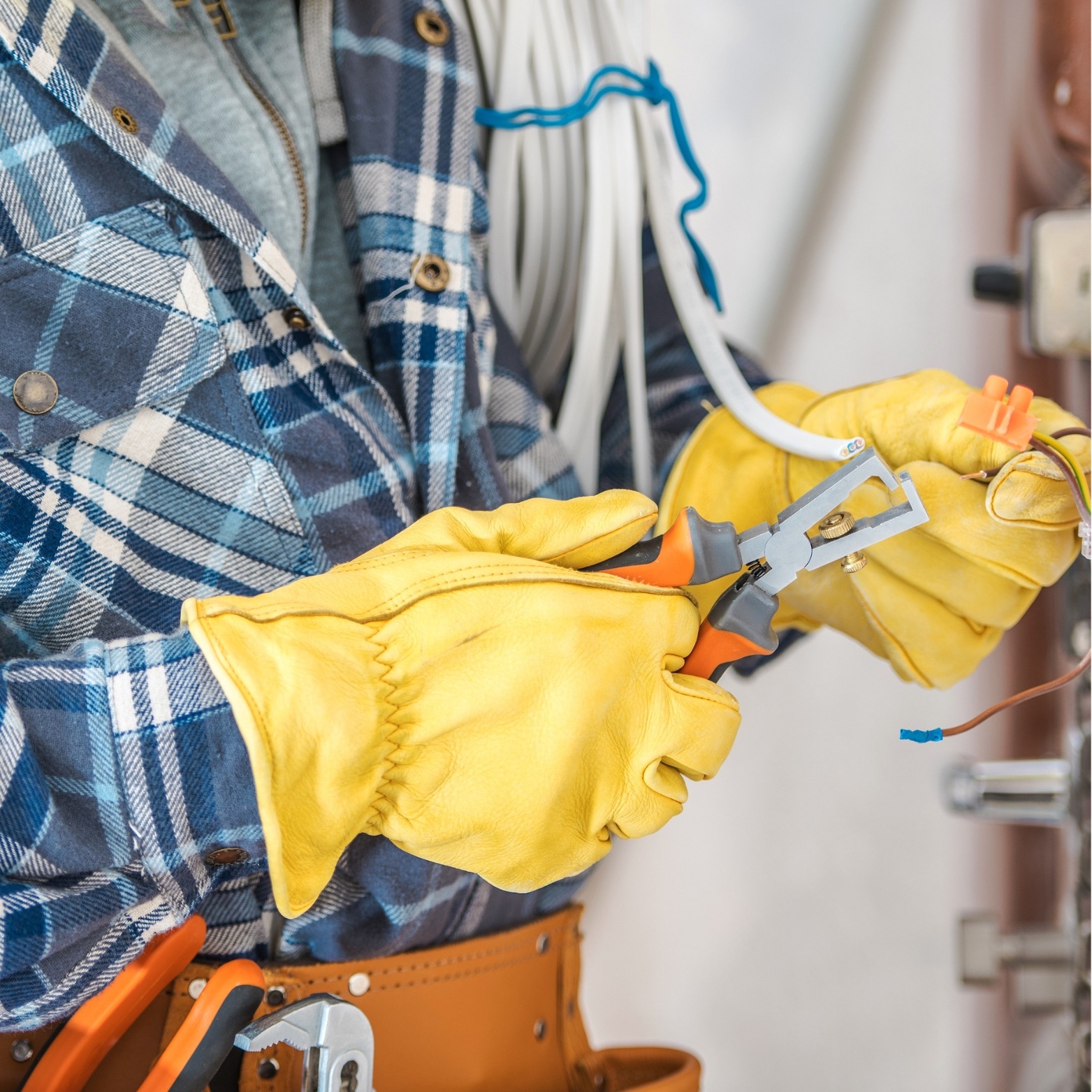 Elektryk w żółtych rękawicach używa ściągacza izolacji na przewodzie, w tle pęk kabli i narzędzia w pasie.