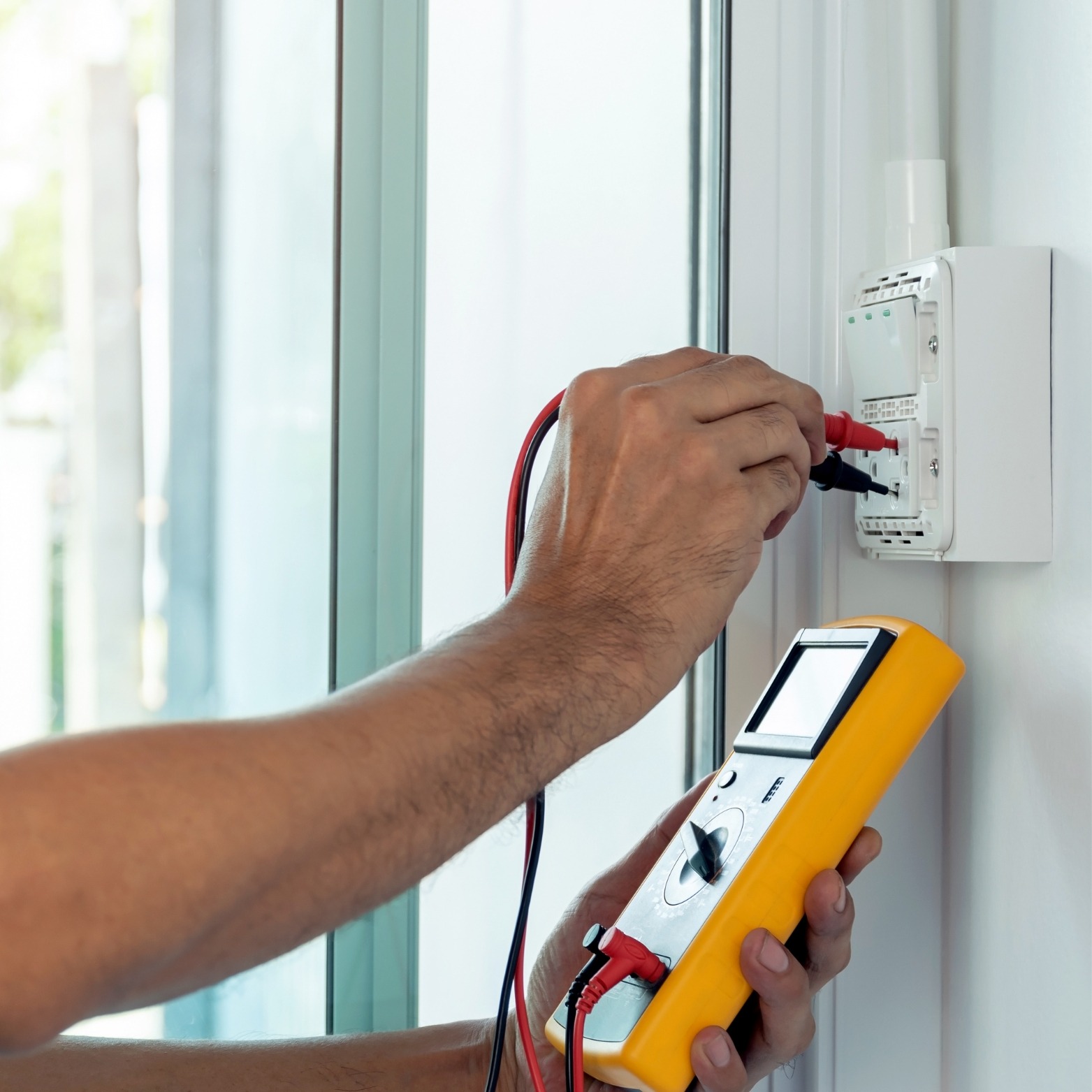 Elektryk mierzy napięcie w gniazdku za pomocą żółtego multimetru, widoczne czerwone i czarne przewody pomiarowe.