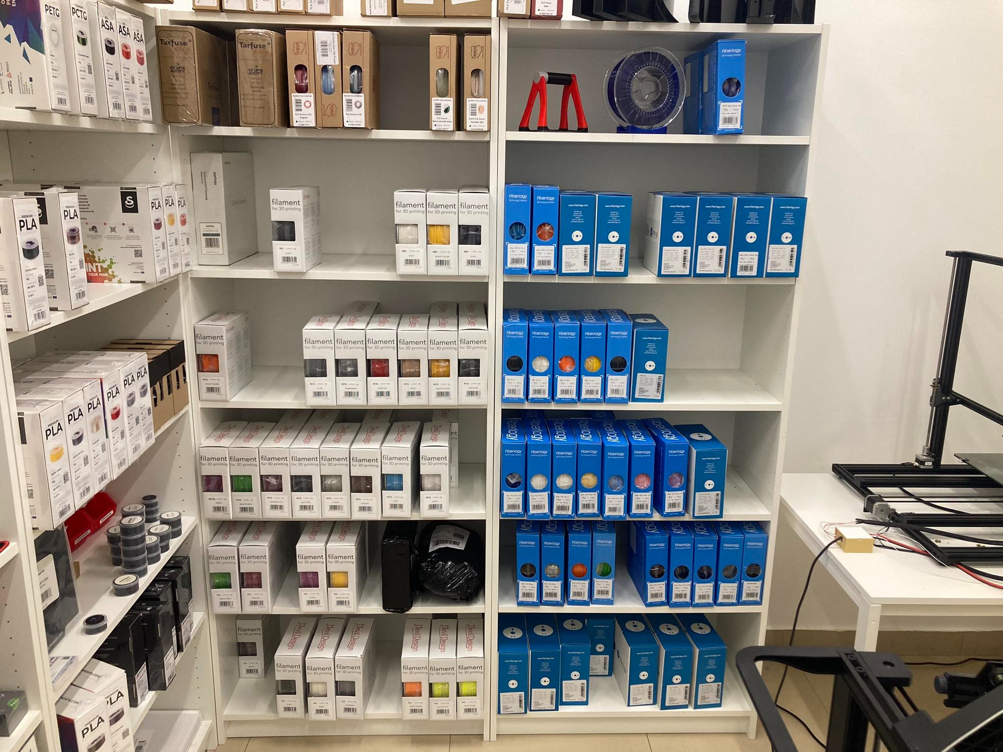 Różnokolorowe szpule filamentów do drukarek 3D, różnych producentów, ułożone rzędami na białych półkach w sklepie.