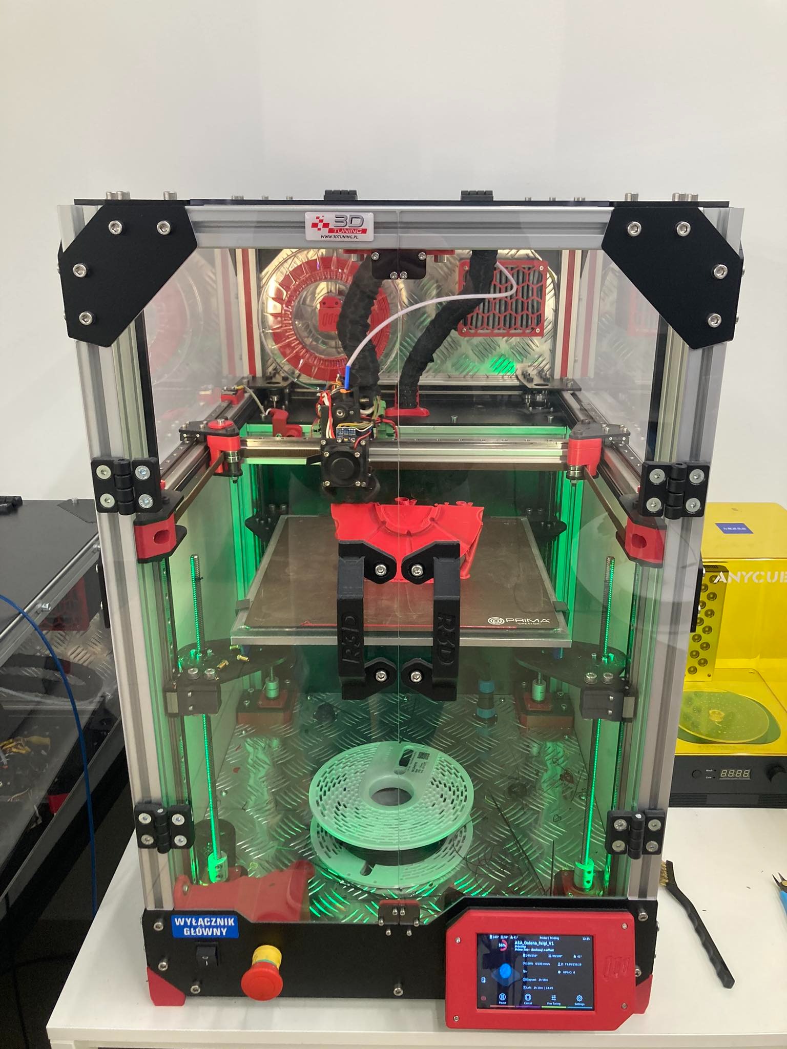 Drukarka 3D z obudową z przezroczystego tworzywa, drukująca czerwony element, z widoczną szpulą filamentu w kolorze miętowym i czerwonym, na metalowej platformie, z panelem sterowania z wyświetlaczem.