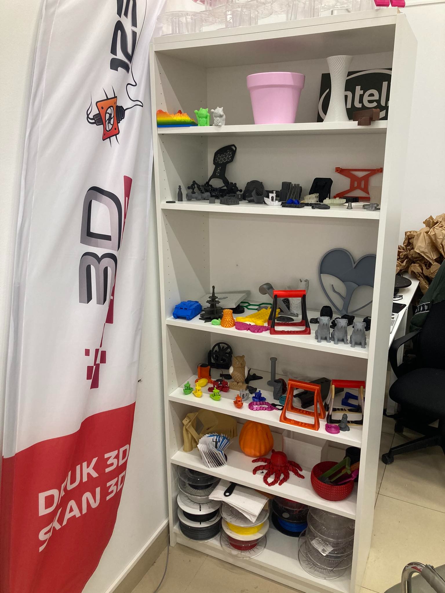 Biały regał z półkami wypełniony różnorodnymi, kolorowymi przedmiotami wydrukowanymi w technologii 3D, obok flaga z napisem 'Druk 3D Skan 3D' i logotypem.