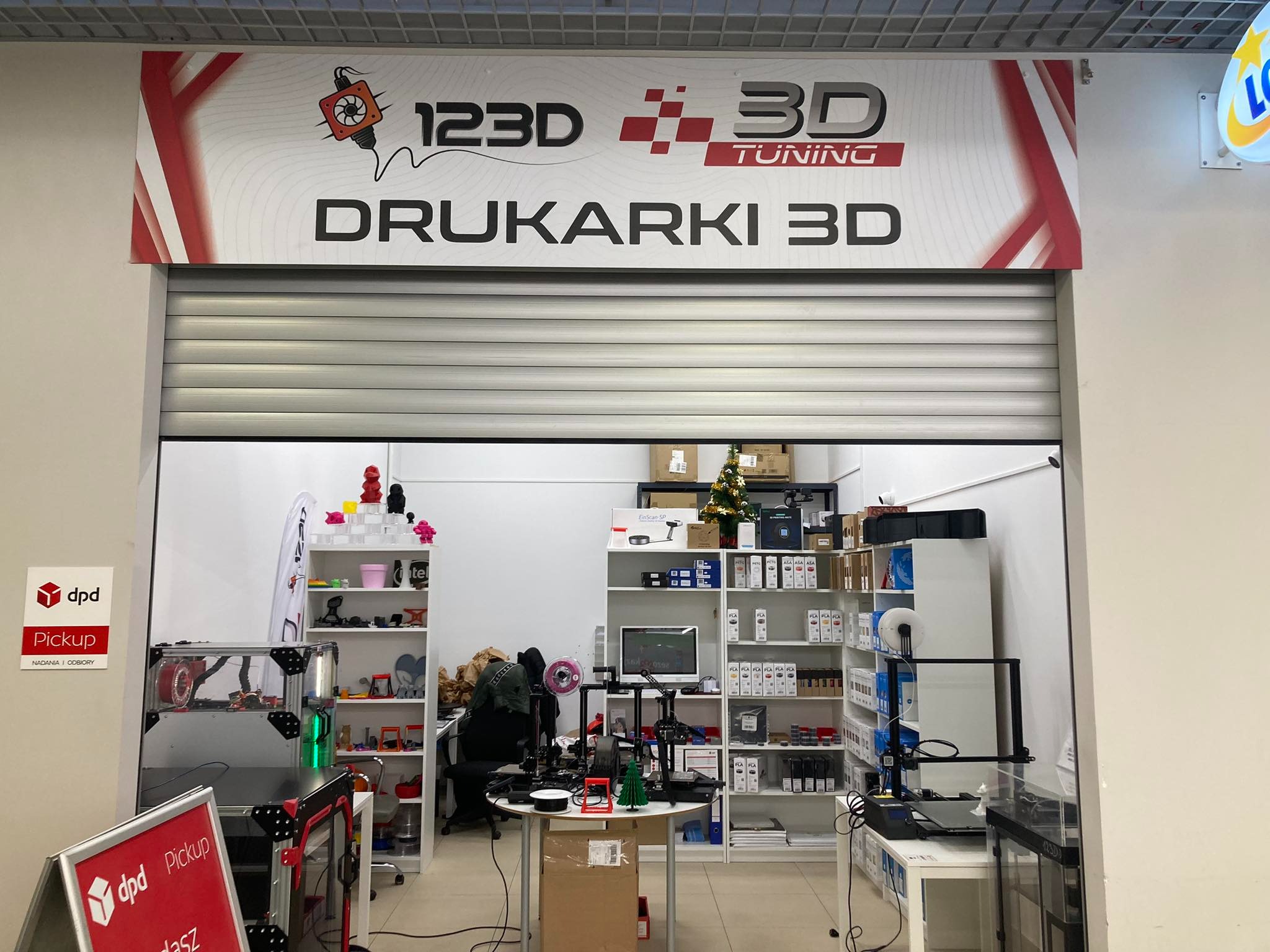 Wnętrze sklepu z drukarkami 3D, widoczne drukarki na stołach i półkach, logo 123D 3D Tuning nad wejściem, punkt nadawania paczek DPD.