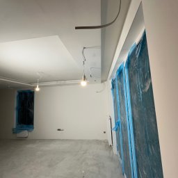 SZYMBUD USŁUGI OGÓLNOBUDOWLANE SZYMON SZYMICHOWSKI - Wnętrze w trakcie prac wykończeniowych: ściany przygotowane do malowania, okna zabezpieczone folią, widoczne kable i tymczasowe oświetlenie.