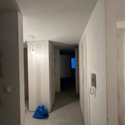 SZYMBUD USŁUGI OGÓLNOBUDOWLANE SZYMON SZYMICHOWSKI - Korytarz w trakcie prac wykończeniowych; ściany z widocznymi śladami po nałożonej gładzi, surowa żarówka wisząca na kablu, niebieski worek na podłodze, szare podłoże, domofon na ścianie.