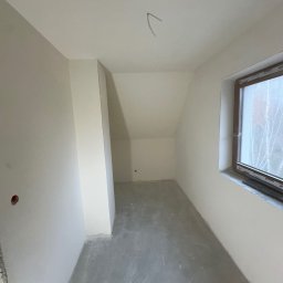 SZYMBUD USŁUGI OGÓLNOBUDOWLANE SZYMON SZYMICHOWSKI - Pokój na poddaszu w trakcie prac wykończeniowych: ściany wstępnie przygotowane, surowe wykończenie podłogi, widoczne otwory instalacyjne w ścianie, okno z widokiem na drzewa.