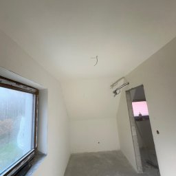 SZYMBUD USŁUGI OGÓLNOBUDOWLANE SZYMON SZYMICHOWSKI - Poddasze po pracach wykończeniowych, świeżo pomalowane ściany na biało, widoczne przewody elektryczne wystające z sufitu, okno z widokiem na zewnątrz, surowa wylewka na podłodze.