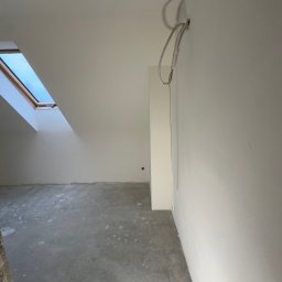 SZYMBUD USŁUGI OGÓLNOBUDOWLANE SZYMON SZYMICHOWSKI - Surowe wnętrze poddasza z oknem dachowym i kablem elektrycznym wychodzącym ze ściany, szara wylewka na podłodze, przygotowane do prac wykończeniowych.