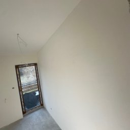 SZYMBUD USŁUGI OGÓLNOBUDOWLANE SZYMON SZYMICHOWSKI - Wnętrze pokoju po nałożeniu gładzi, widoczne gładkie ściany i sufit, surowy beton na podłodze, okno z widokiem na zewnątrz, wystające przewody elektryczne.