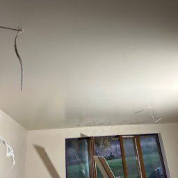 SZYMBUD USŁUGI OGÓLNOBUDOWLANE SZYMON SZYMICHOWSKI - Świeżo położona, jasna gładź na suficie z wystającymi przewodami elektrycznymi, okno zabezpieczone taśmą malarską, widoczne płyty karton-gips oparte o okno.