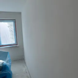 Wnętrze pokoju po nałożeniu gładzi, okno zabezpieczone folią, widoczny fragment podłogi z pozostałościami po pracach.