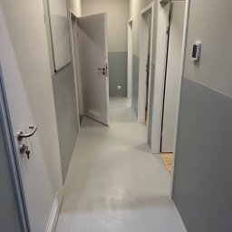 SZYMBUD USŁUGI OGÓLNOBUDOWLANE SZYMON SZYMICHOWSKI - Szary korytarz biurowy po remoncie, z pomalowanymi ścianami na szaro, jasną podłogą żywiczną, białymi drzwiami z srebrnymi klamkami i wentylacją oraz tablicą suchościeralną.