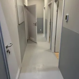 Szary korytarz biurowy po remoncie, z pomalowanymi ścianami na szaro, jasną podłogą żywiczną, białymi drzwiami z srebrnymi klamkami i wentylacją oraz tablicą suchościeralną.