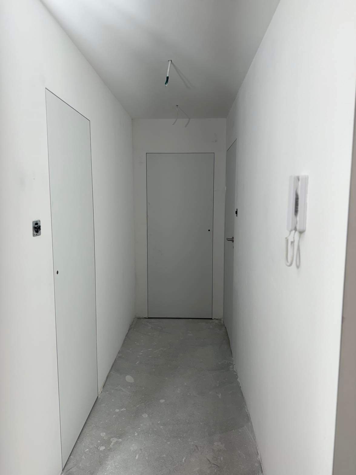 Korytarz w stanie deweloperskim z gładkimi, białymi ścianami, dwiema ukrytymi drzwiami po lewej stronie i domofonem po prawej. Podłoga betonowa, sufit z wystającymi kablami.