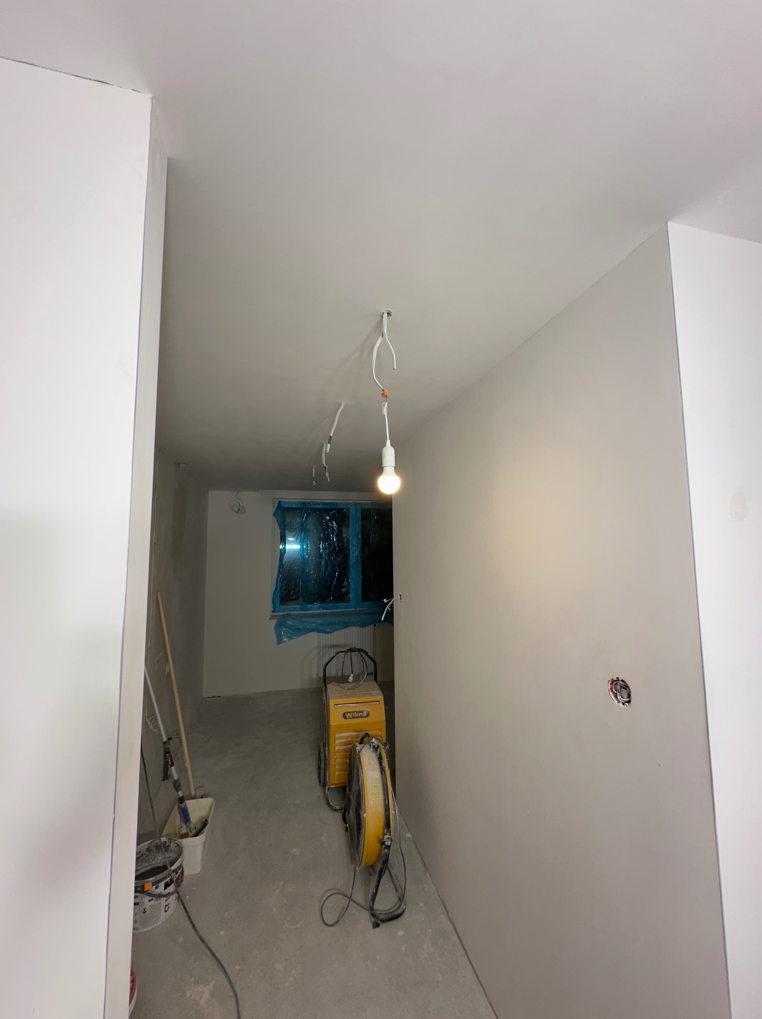 Wnętrze w trakcie prac wykończeniowych: świeżo pomalowane ściany w odcieniach szarości, zabezpieczone okno folią, osuszacz powietrza Wilms, wiadro z narzędziami malarskimi w rogu pomieszczenia...