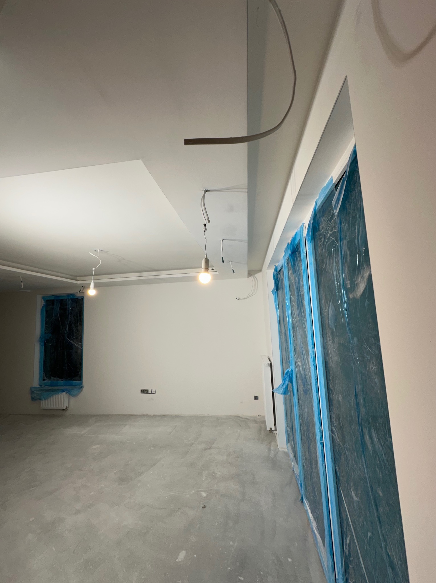 Wnętrze w trakcie prac wykończeniowych: ściany przygotowane do malowania, okna zabezpieczone folią, widoczne kable i tymczasowe oświetlenie.