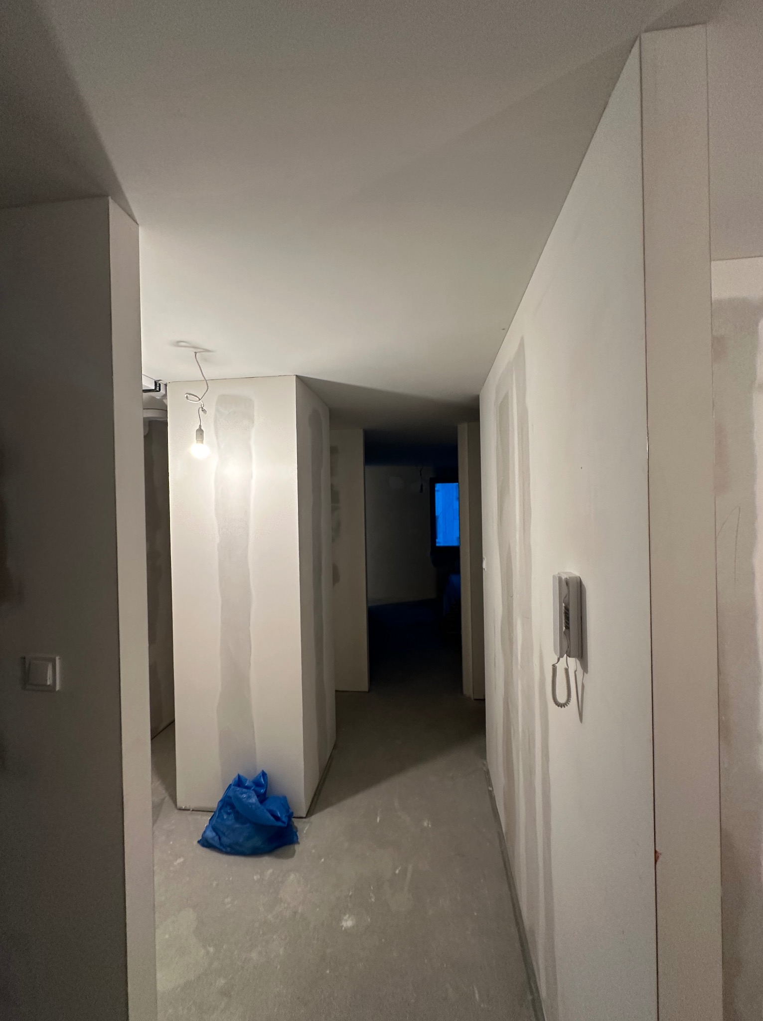 Korytarz w trakcie prac wykończeniowych; ściany z widocznymi śladami po nałożonej gładzi, surowa żarówka wisząca na kablu, niebieski worek na podłodze, szare podłoże, domofon na ścianie.