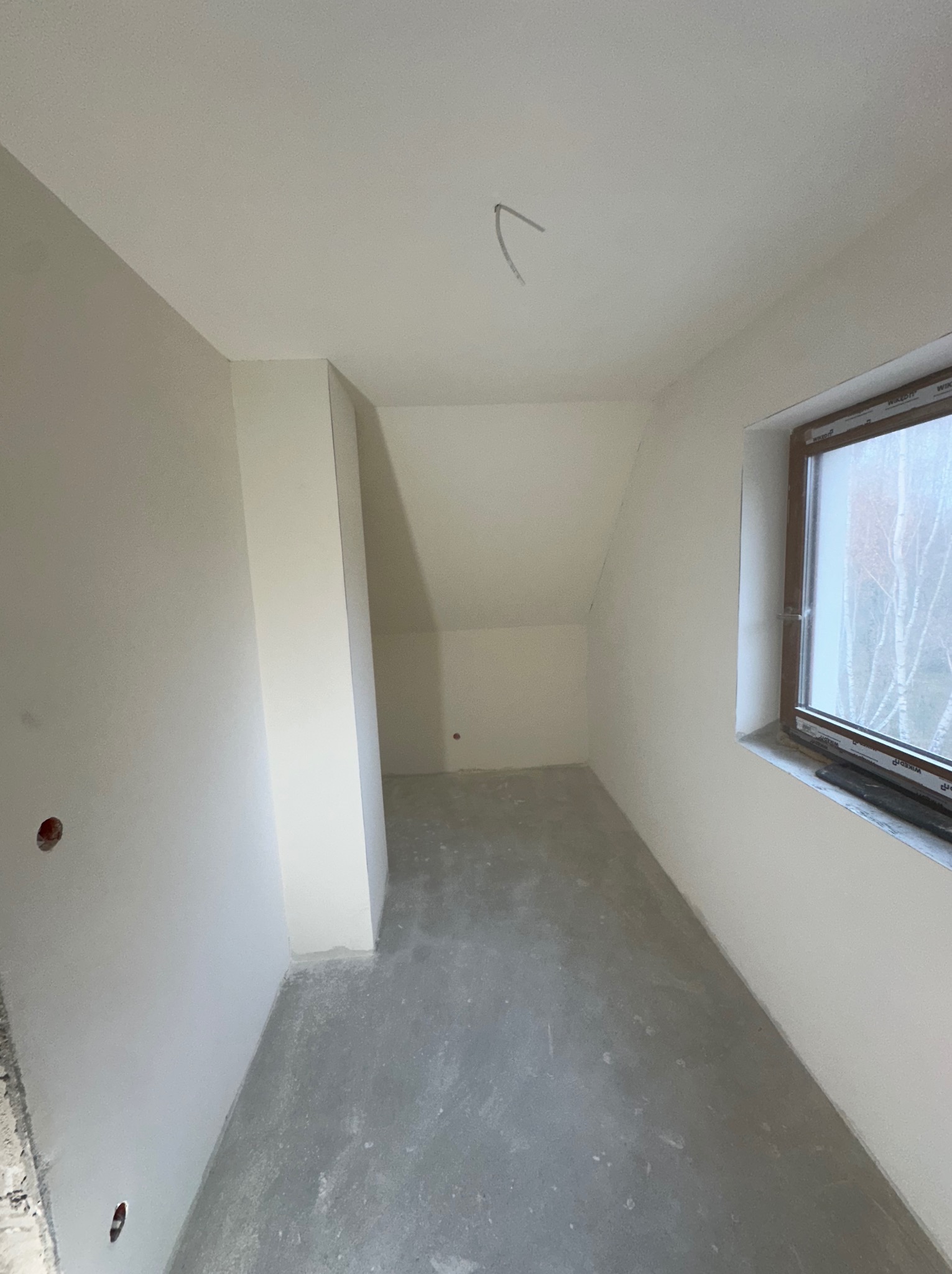 Pokój na poddaszu w trakcie prac wykończeniowych: ściany wstępnie przygotowane, surowe wykończenie podłogi, widoczne otwory instalacyjne w ścianie, okno z widokiem na drzewa.