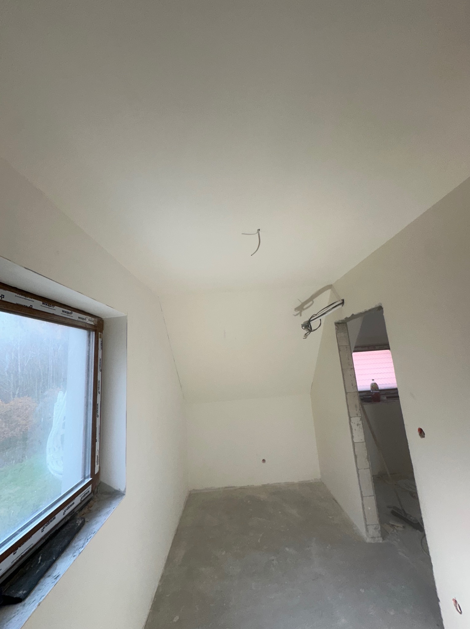 Poddasze po pracach wykończeniowych, świeżo pomalowane ściany na biało, widoczne przewody elektryczne wystające z sufitu, okno z widokiem na zewnątrz, surowa wylewka na podłodze.