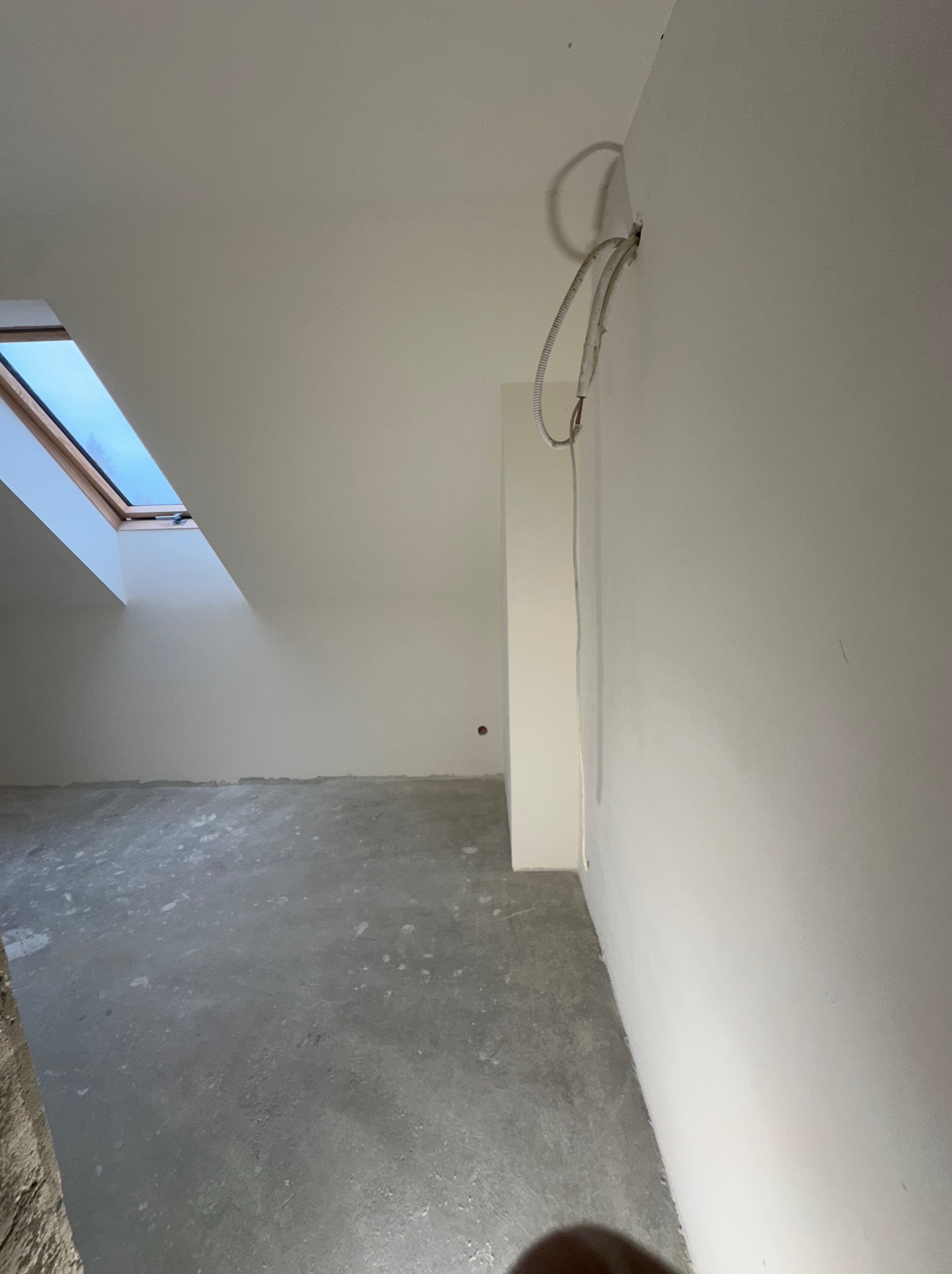 Surowe wnętrze poddasza z oknem dachowym i kablem elektrycznym wychodzącym ze ściany, szara wylewka na podłodze, przygotowane do prac wykończeniowych.