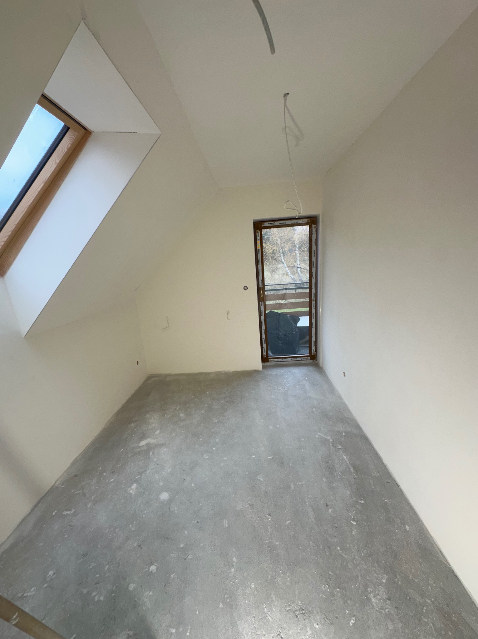 Poddasze po remoncie: gładkie, pomalowane ściany w jasnym odcieniu, surowa betonowa podłoga, okno dachowe i balkonowe z widokiem na drzewa.