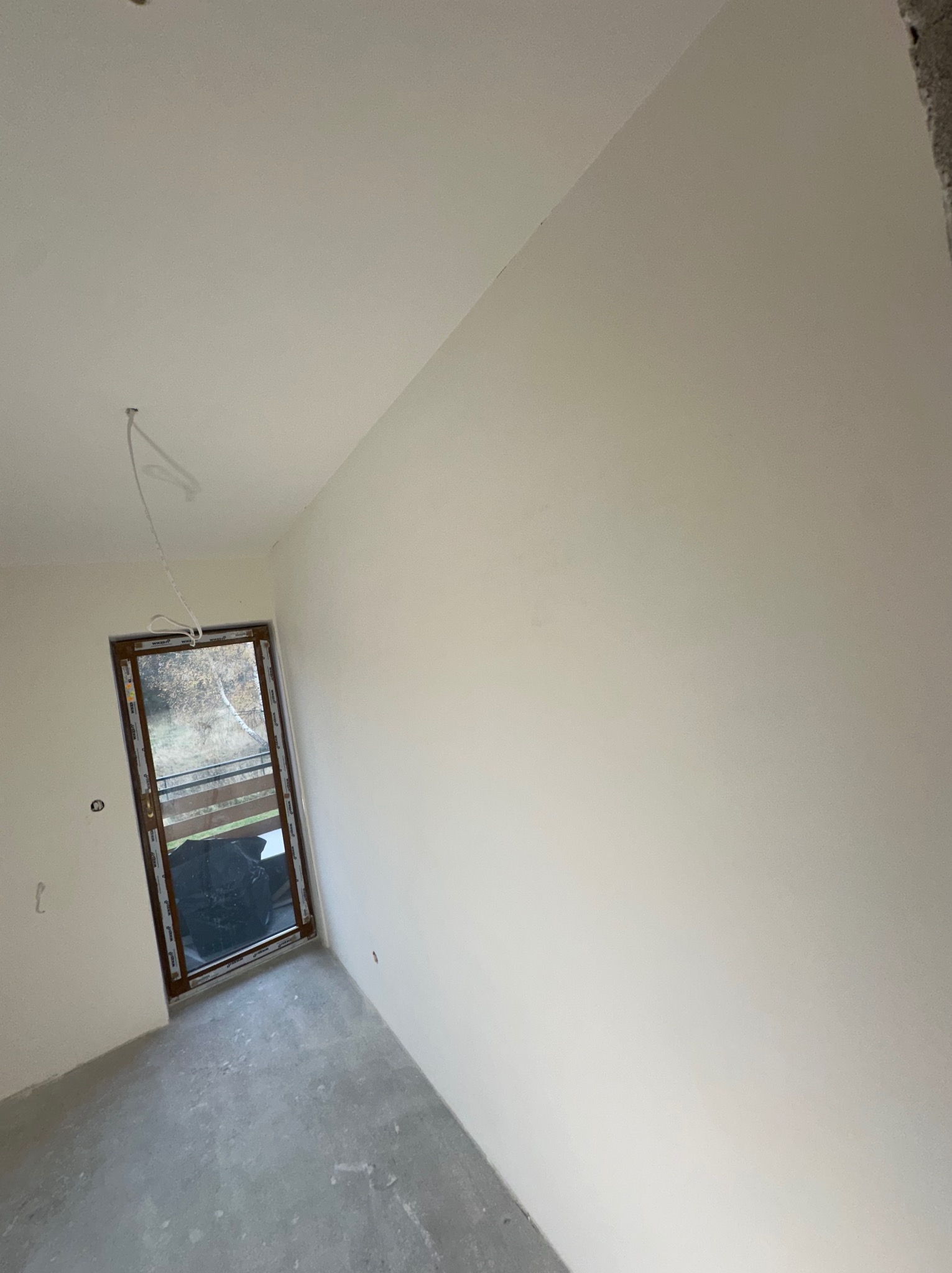 Wnętrze pokoju po nałożeniu gładzi, widoczne gładkie ściany i sufit, surowy beton na podłodze, okno z widokiem na zewnątrz, wystające przewody elektryczne.