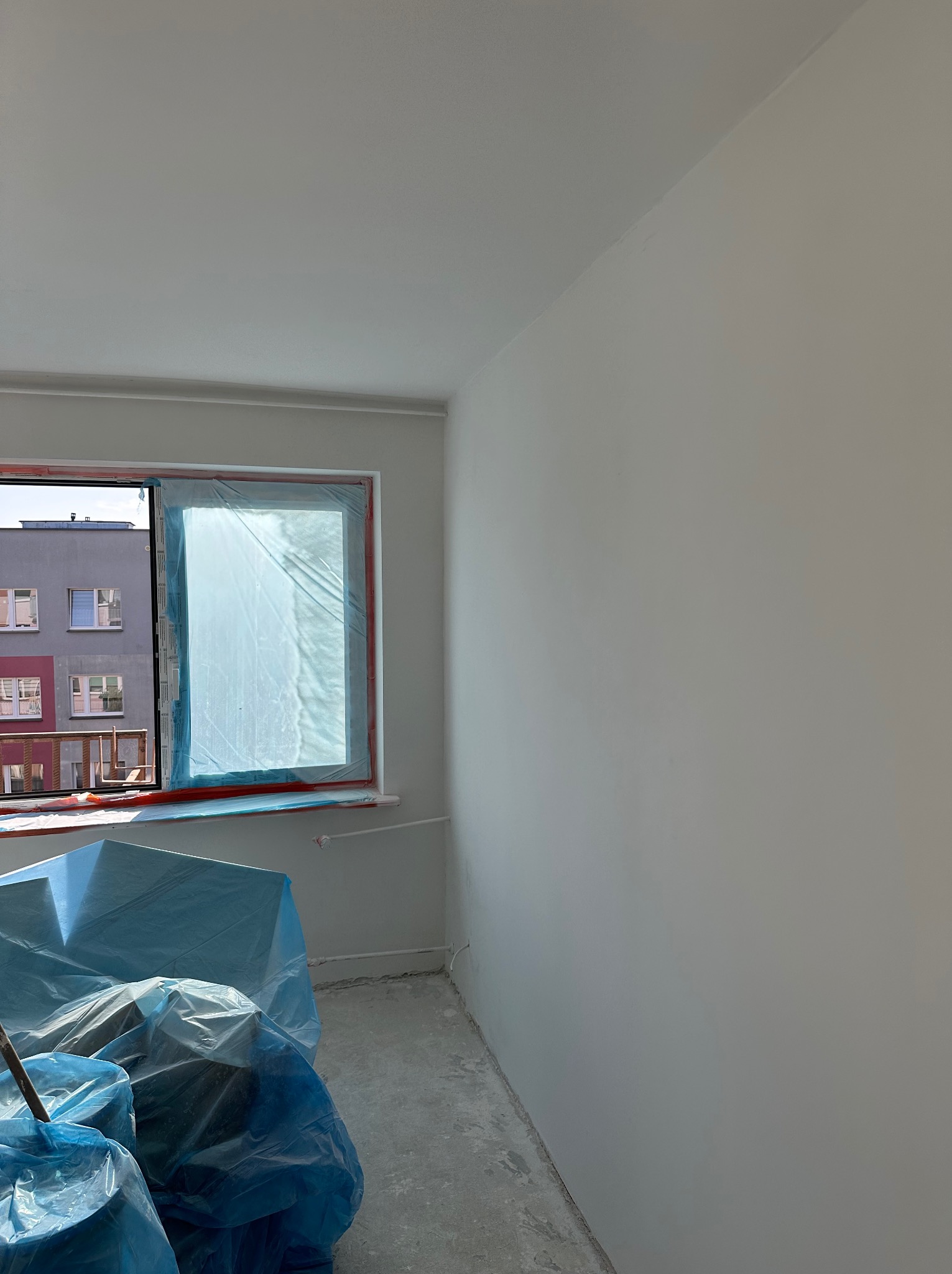 Świeżo pomalowane na biało ściany w pustym pokoju, okno zabezpieczone folią malarską, podłoga częściowo zakryta folią ochronną, widoczne wiaderka z farbą.
