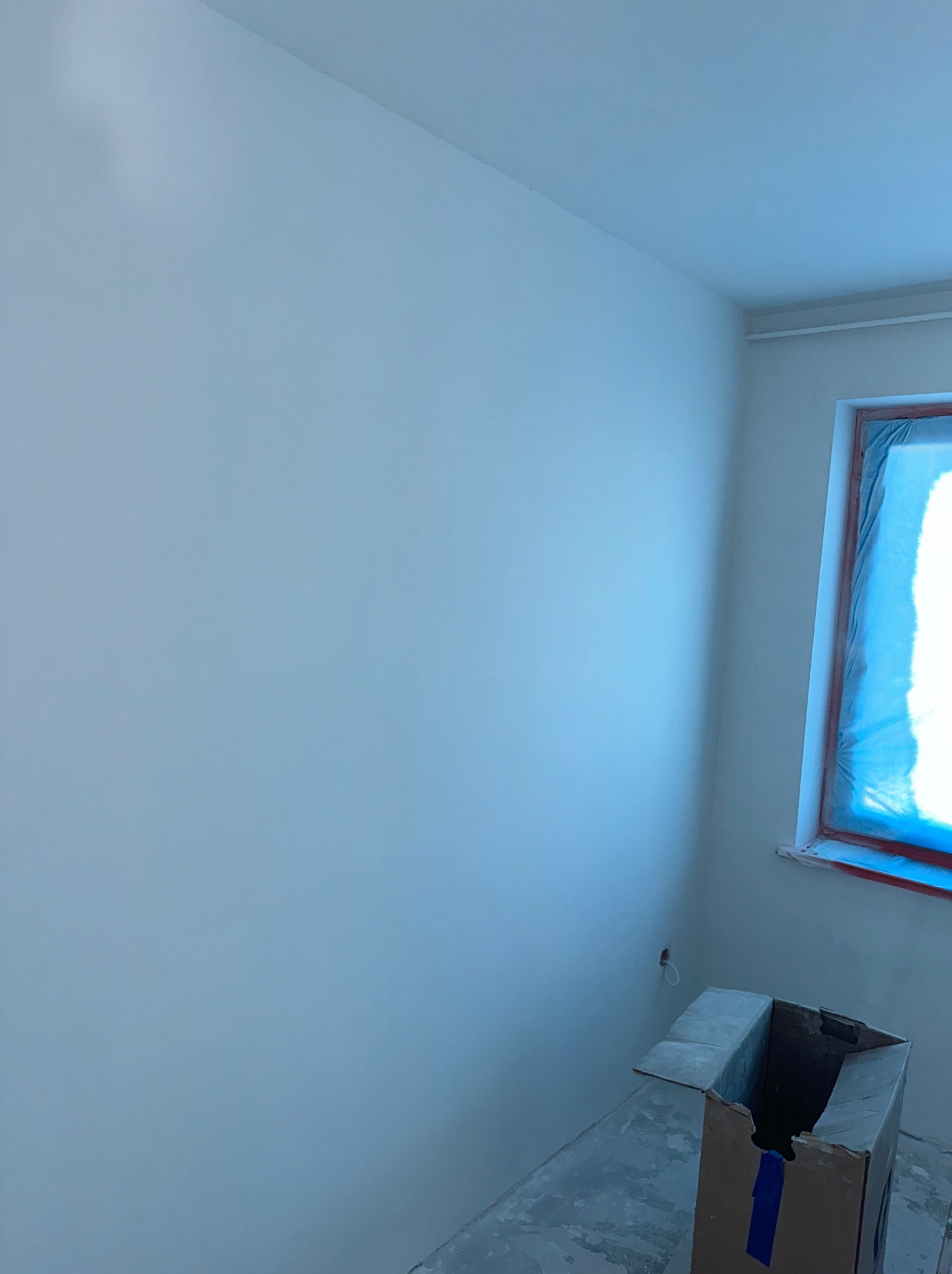Świeżo pomalowane, jasne wnętrze z oknem zabezpieczonym folią malarską, karton na podłodze, widoczny fragment gładkiej ściany.