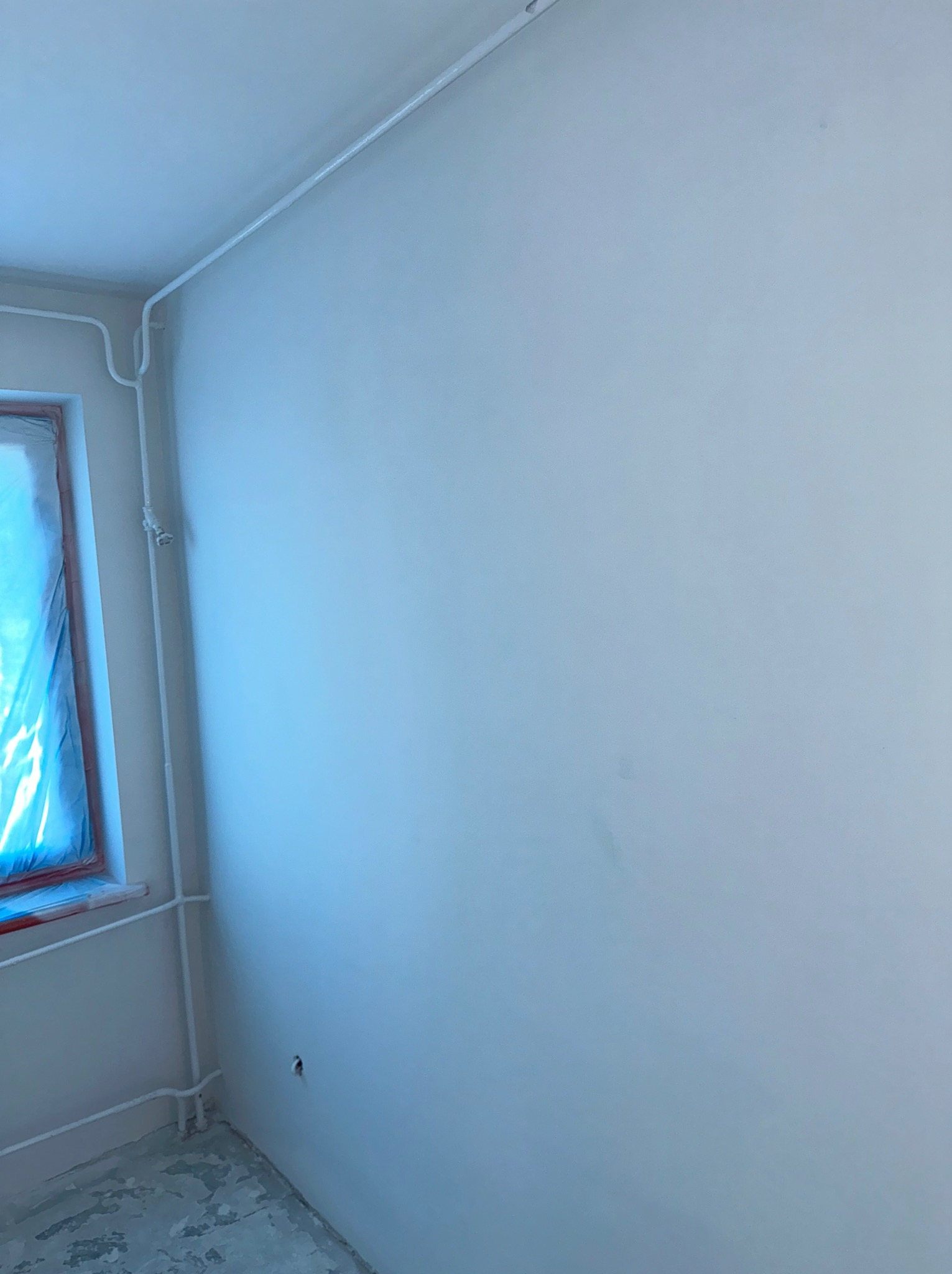 Gładka, biała ściana po remoncie, z widocznymi przewodami elektrycznymi w listwach przypodłogowych i oknem zabezpieczonym folią malarską.