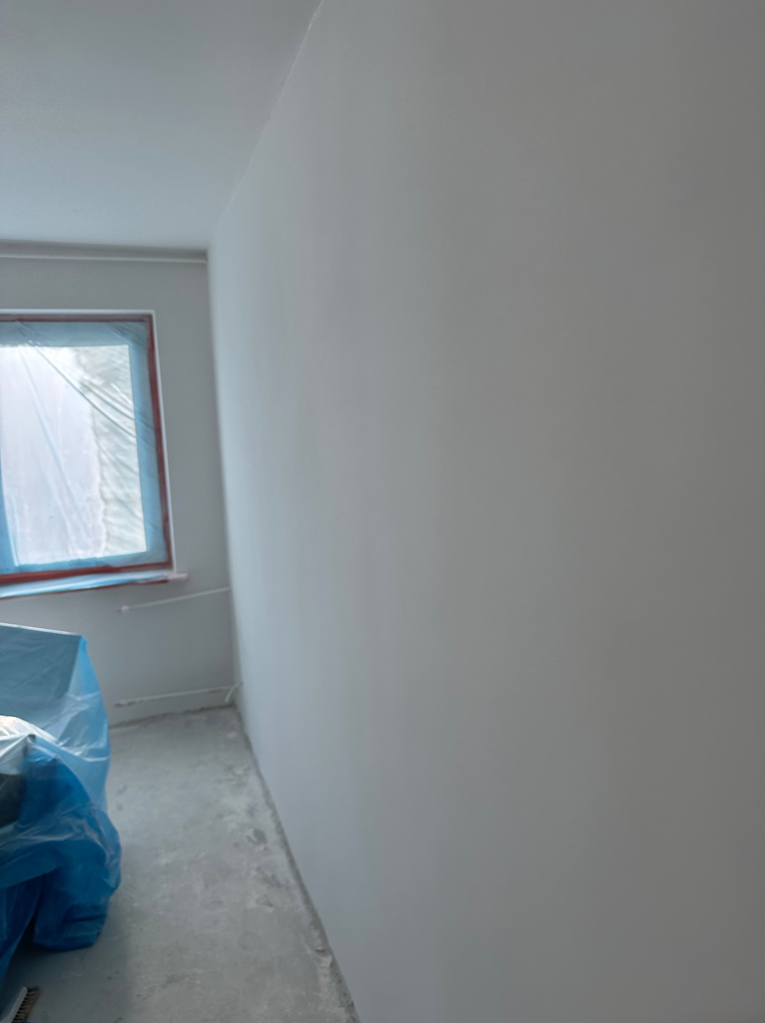 Wnętrze pokoju po nałożeniu gładzi, okno zabezpieczone folią, widoczny fragment podłogi z pozostałościami po pracach.