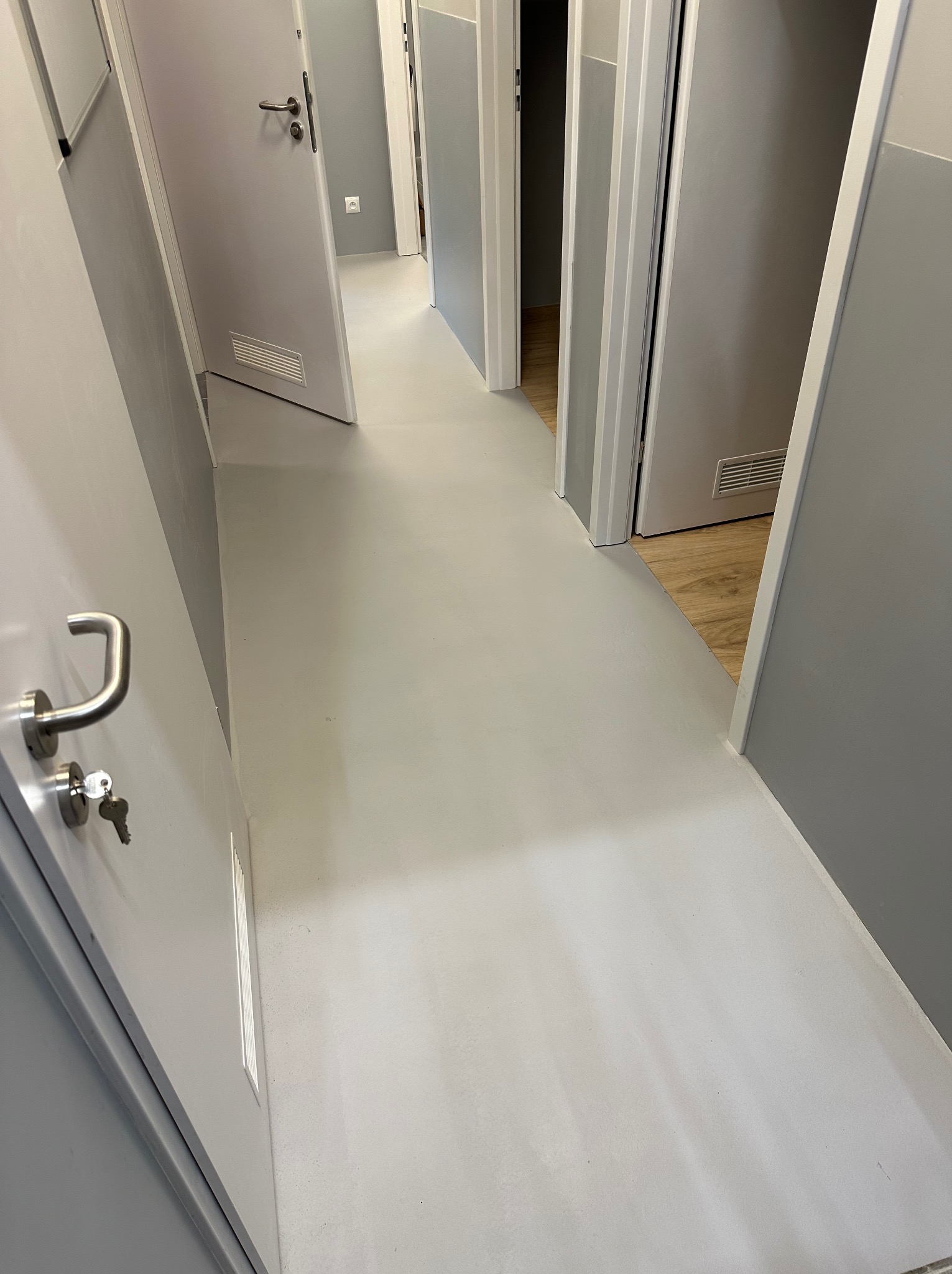 Korytarz po remoncie z szarą podłogą z żywicy epoksydowej, widoczne drzwi z metalowymi klamkami i kluczami w zamku, w tle fragment podłogi wyłożonej panelami.