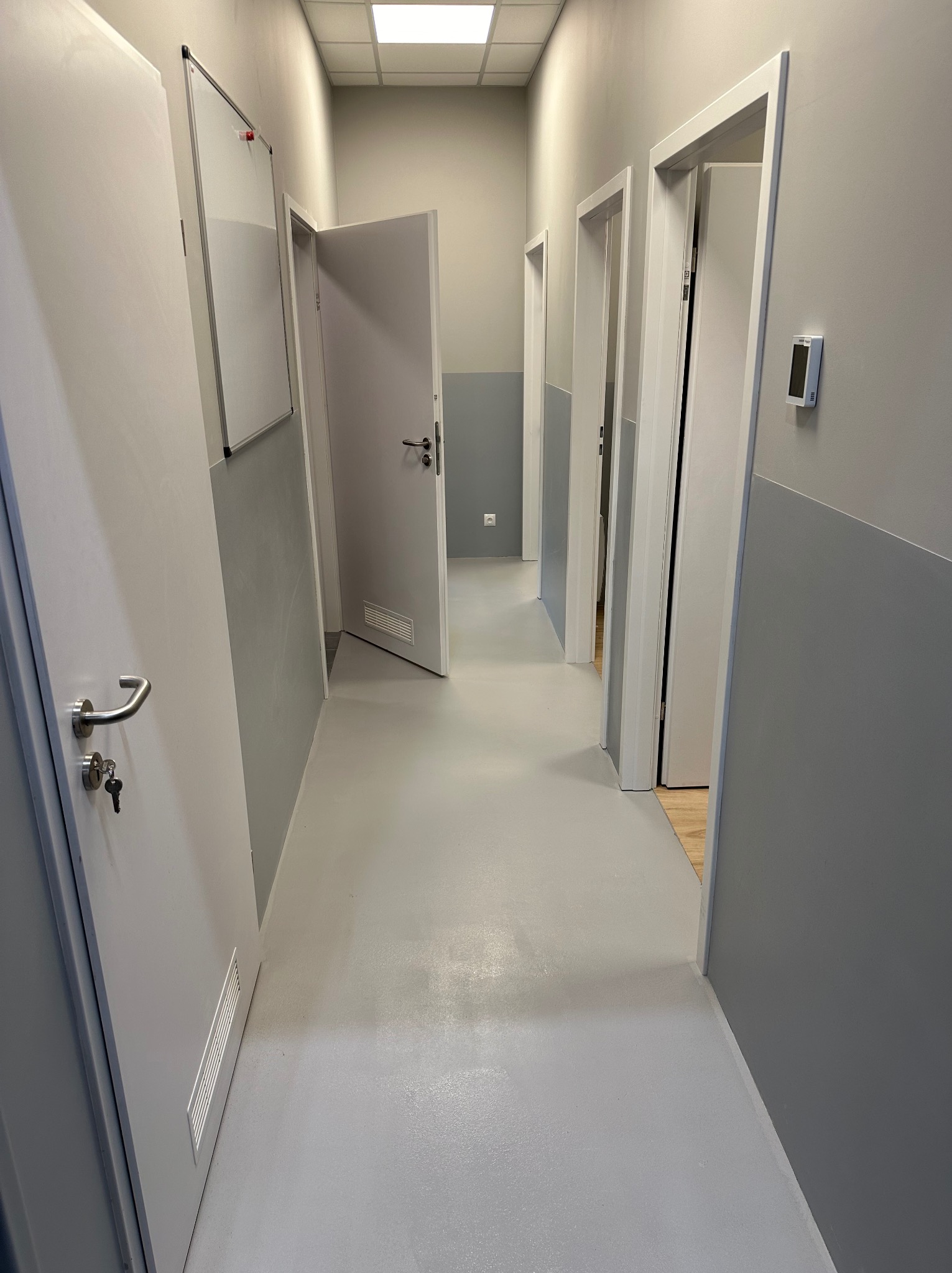 Szary korytarz biurowy po remoncie, z pomalowanymi ścianami na szaro, jasną podłogą żywiczną, białymi drzwiami z srebrnymi klamkami i wentylacją oraz tablicą suchościeralną.