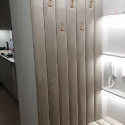 Beżowy panel tapicerowany z siedziskiem i złotymi haczykami w przedpokoju, oświetlenie LED na górze, widoczne ślady użytkowania na panelu.