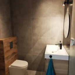 Nowoczesna łazienka z grafitowymi płytkami, podwieszanym WC z drewnianą obudową, białą umywalką na szafce z drewnianymi frontami i okrągłym lustrem podświetlonym punktowym światłem.