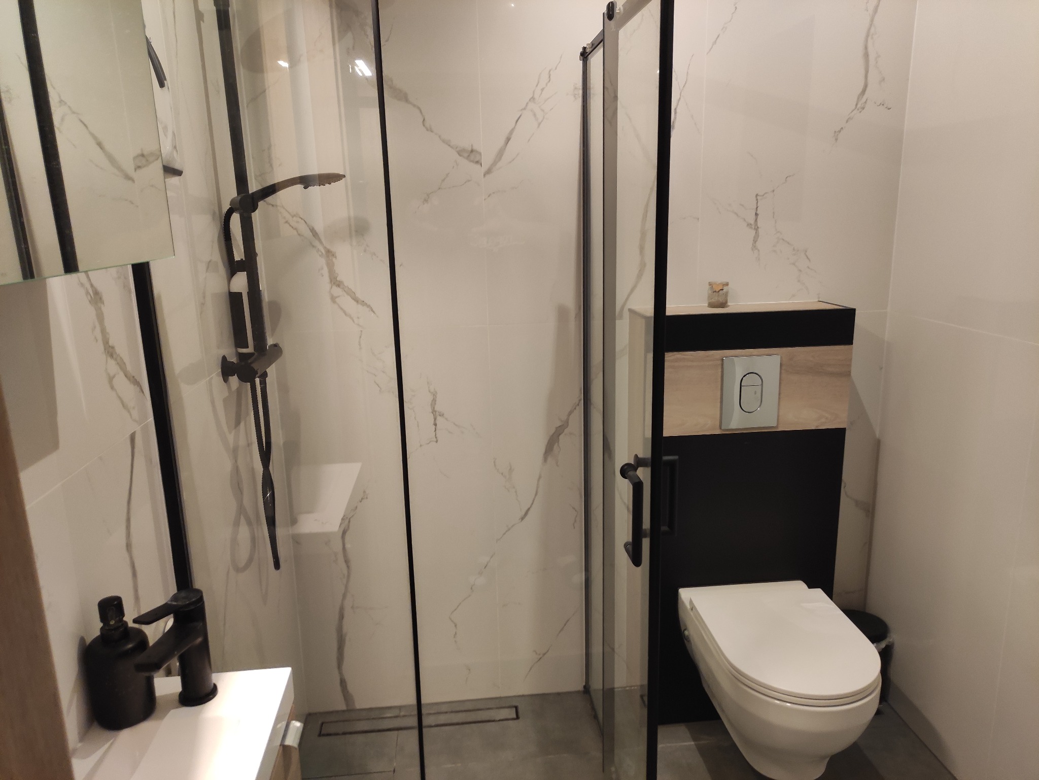 Nowoczesna łazienka z kabiną prysznicową typu walk-in z czarnymi profilami, białą armaturą, marmurowymi płytkami na ścianach i czarną baterią umywalkową.