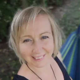 Portret uśmiechniętej blondynki z krótkimi włosami, w czarnym topie, z delikatnym makijażem oczu, na tle zieleni i fragmentu niebiesko-zielonego hamaka.