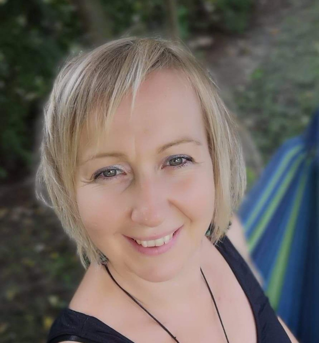 Portret uśmiechniętej blondynki z krótkimi włosami, w czarnym topie, z delikatnym makijażem oczu, na tle zieleni i fragmentu niebiesko-zielonego hamaka.