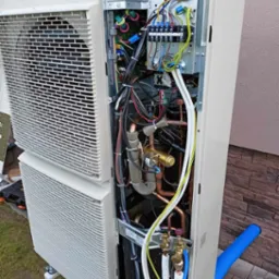 Otwarta obudowa zewnętrznej jednostki klimatyzacji z widocznymi przewodami elektrycznymi, miedzianymi rurami i zaworami, na tle ściany budynku.