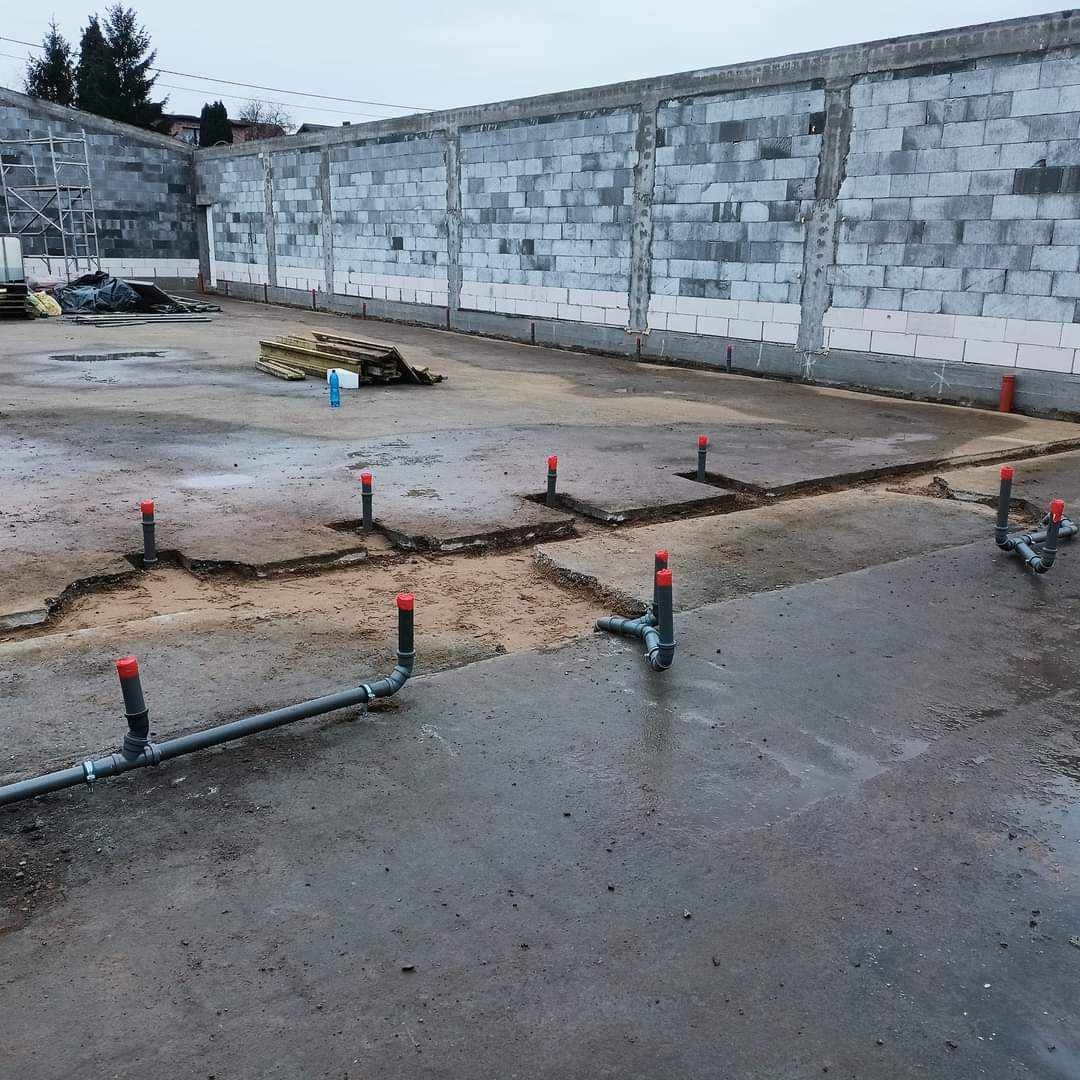 Montaż instalacji kanalizacyjnej w posadzce budynku w Milejowie, widoczne rury z czerwonymi zaślepkami wystające z wykopów w betonie, szara ściana z bloczków w tle.