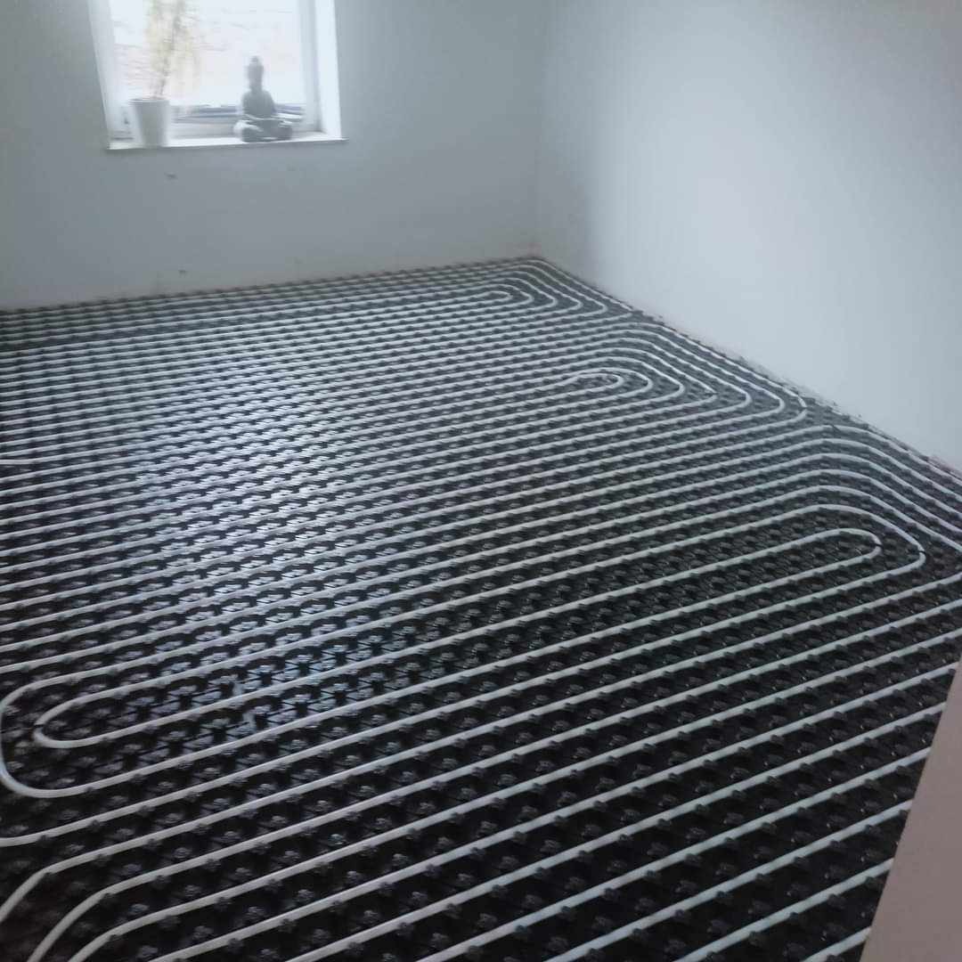 Białe rurki ogrzewania podłogowego ułożone w pętle na czarnej macie izolacyjnej w jasnym pomieszczeniu, widok z góry na instalację przed zalaniem wylewką.