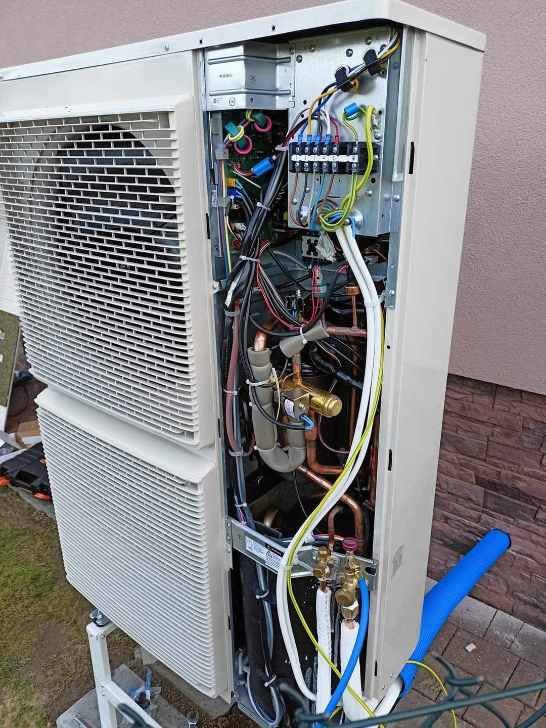 Otwarta obudowa zewnętrznej jednostki klimatyzacji z widocznymi przewodami elektrycznymi, miedzianymi rurami i zaworami, na tle ściany budynku.