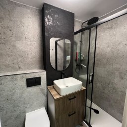 Don_Budowa - Nowoczesna łazienka z szarymi płytkami, kabiną prysznicową z czarnymi ramami, umywalką nablatową na szafce z drewnopodobnymi frontami i owalnym lustrem w czarnej ramie.