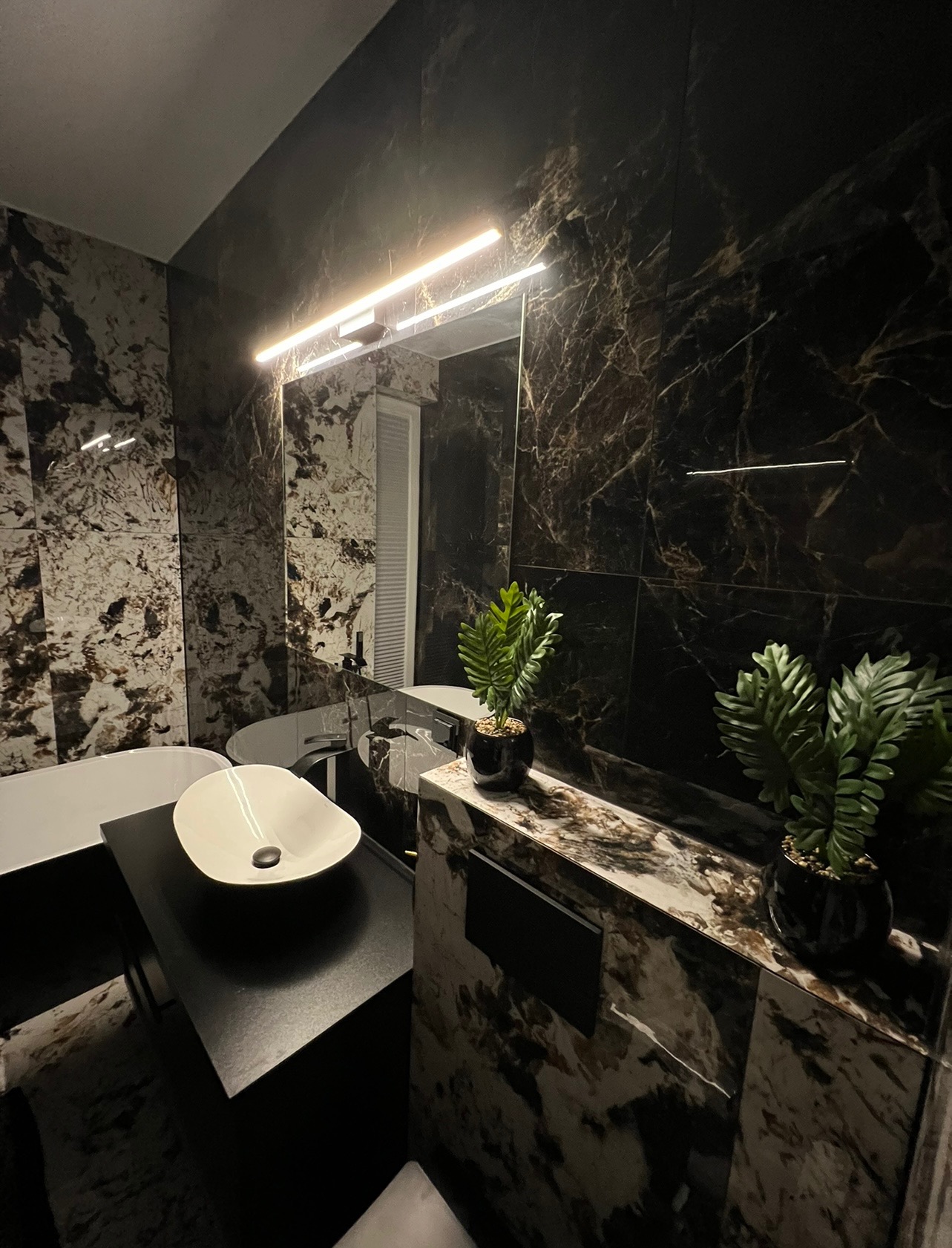 Nowoczesna łazienka z czarnymi marmurkowymi płytkami, owalną białą umywalką nablatową, czarną wanną wolnostojącą i podświetlanym lustrem.