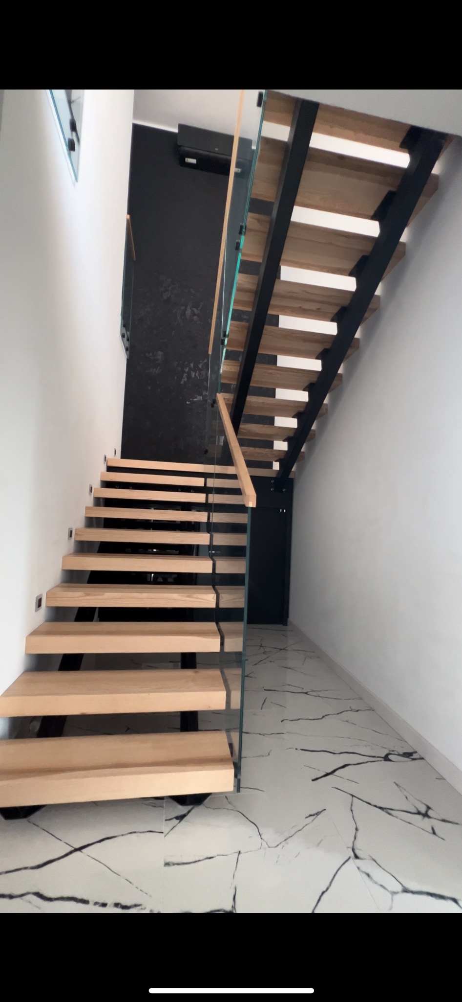 Nowoczesne schody drewniane z czarną metalową konstrukcją i szklaną balustradą, prowadzące na czarne piętro, wykończone białą ścianą i marmurową podłogą.