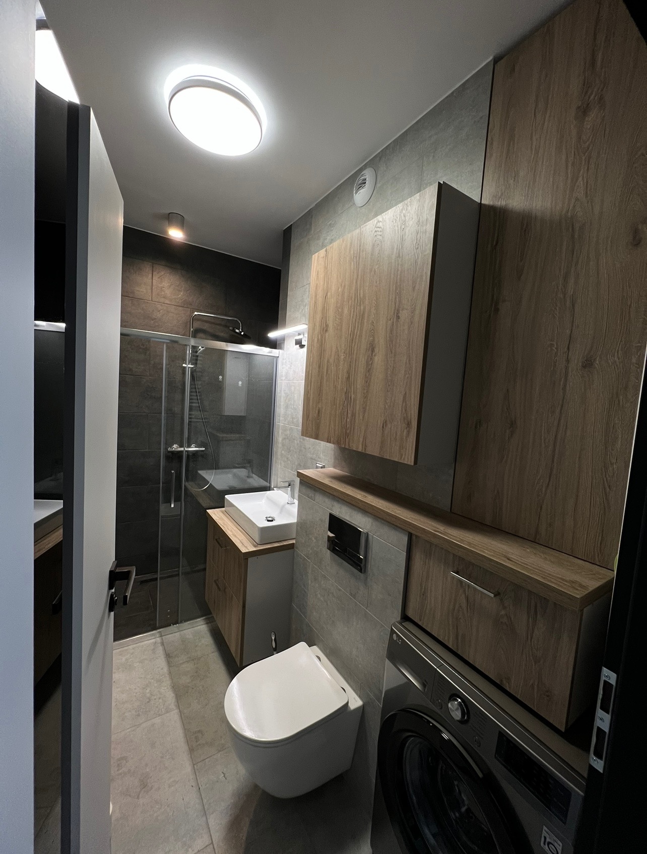 Nowoczesna łazienka z kabiną prysznicową, umywalką nablatową, toaletą wiszącą i zabudową meblową w odcieniach szarości i drewna, oświetlona okrągłą lampą sufitową.