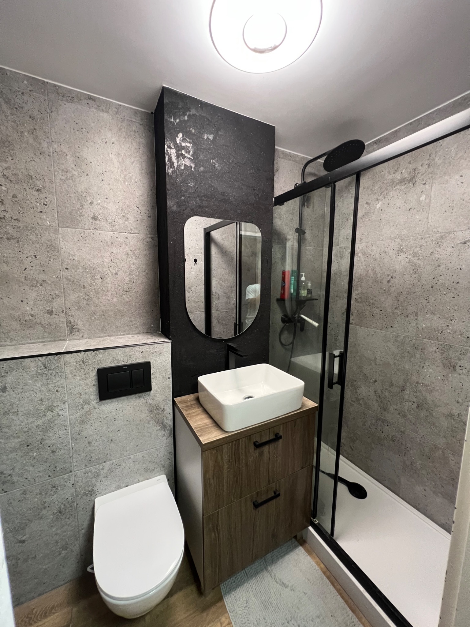 Nowoczesna łazienka z szarymi płytkami, kabiną prysznicową z czarnymi ramami, umywalką nablatową na szafce z drewnopodobnymi frontami i owalnym lustrem w czarnej ramie.