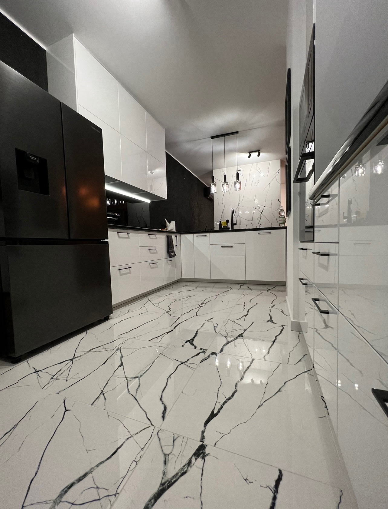 Nowoczesna kuchnia z białymi, lakierowanymi szafkami, czarnymi blatami i podłogą z białych płytek imitujących marmur z czarnymi żyłami. Widoczna czarna lodówka i czarne dodatki.