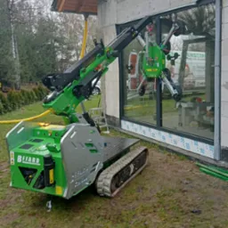 Montaż dużego okna z użyciem zielonego podnośnika gąsienicowego marki Befard na placu budowy. Na szybie widoczne przyssawki podnośnika. W tle budynek w stanie surowym i częściowo widoczny samochód...