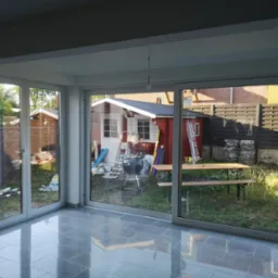 Nowoczesne, przeszklone drzwi przesuwne z widokiem na ogród, gdzie widać altankę, grill i stół piknikowy. Podłoga z szarych płytek.