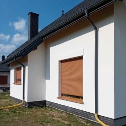 Wojciech Besser Zoom na Dom - Świeżo pomalowana, biała elewacja domu jednorodzinnego z brązowymi roletami zewnętrznymi i czarnymi elementami dachu oraz ceglaną podmurówką, widok z boku, wąż odprowadzający wodę leży na trawie.