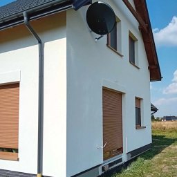 Wojciech Besser Zoom na Dom - Świeżo pomalowana, biała elewacja domu jednorodzinnego z brązowymi roletami i anteną satelitarną na ścianie, widok z zewnątrz w słoneczny dzień, z widocznym fragmentem dachu i rynny.